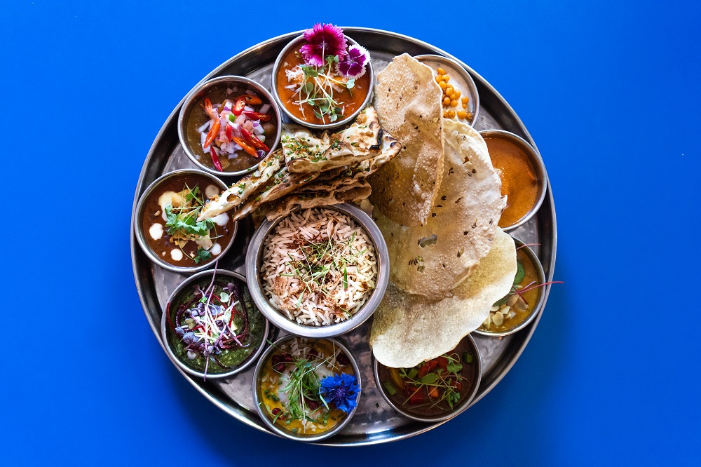 A vibrant Indian thali set against a blue background, showcasing a variety of dishes including curries, rice, and papad, garnished with flowers and sprouts.