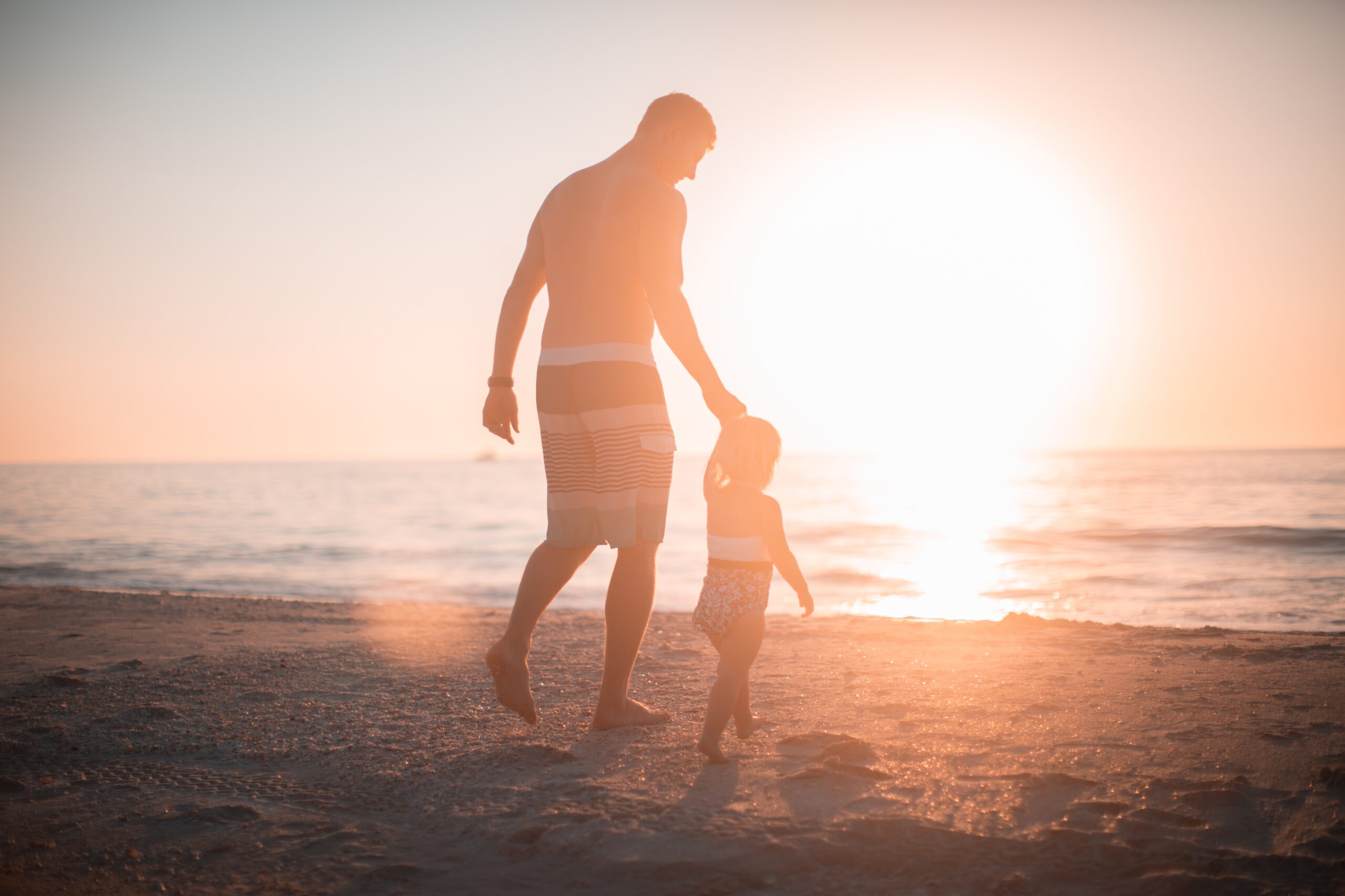 A man and a young child walking hand in hand on a sandy beach during sunset, with the sun casting a warm golden glow over the scene. The ocean waves gently lap at the shore in the background.