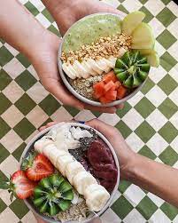 Two hands holding smoothie bowls with assorted fruits and toppings at Blended Health Bar, Noosa.