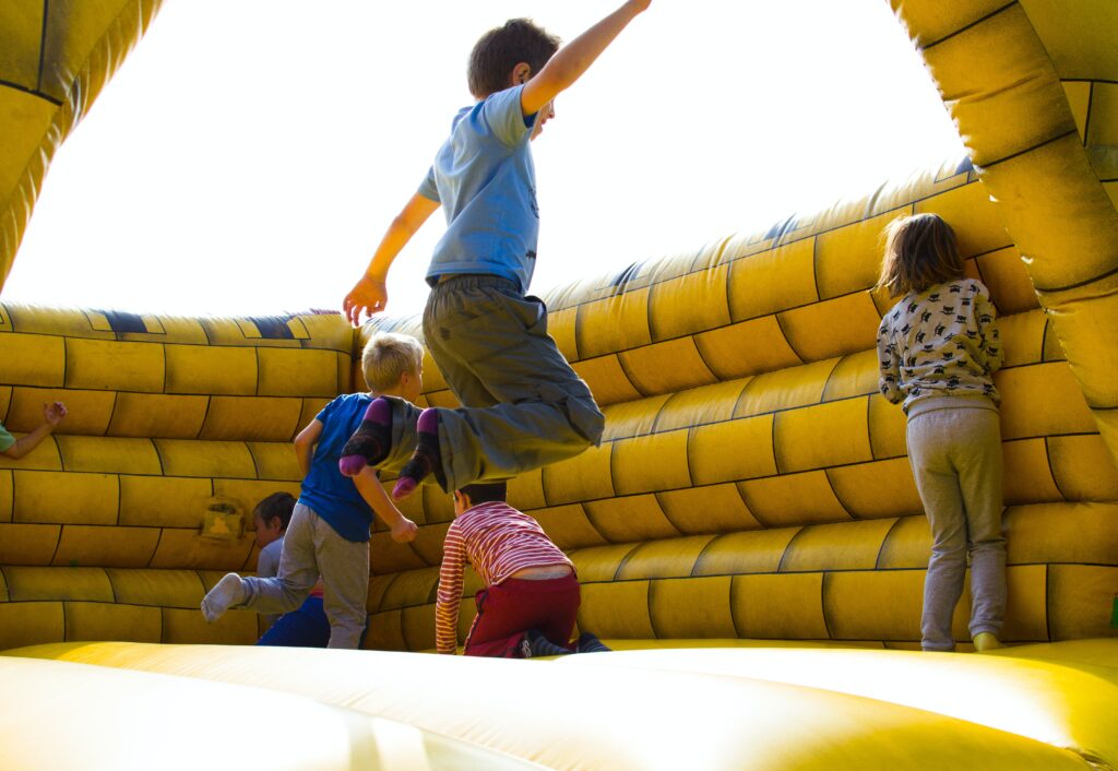 Children playing on a yellow inflatable bounce house under bright sunlight. One child is in mid-air, leaping joyfully, while others are climbing and jumping inside the bouncy structure. A girl, seen from the back, stands on the edge, looking inside. The bounce house's vivid yellow color and the kids' energetic activity convey a sense of fun and liveliness typical of outdoor play areas.