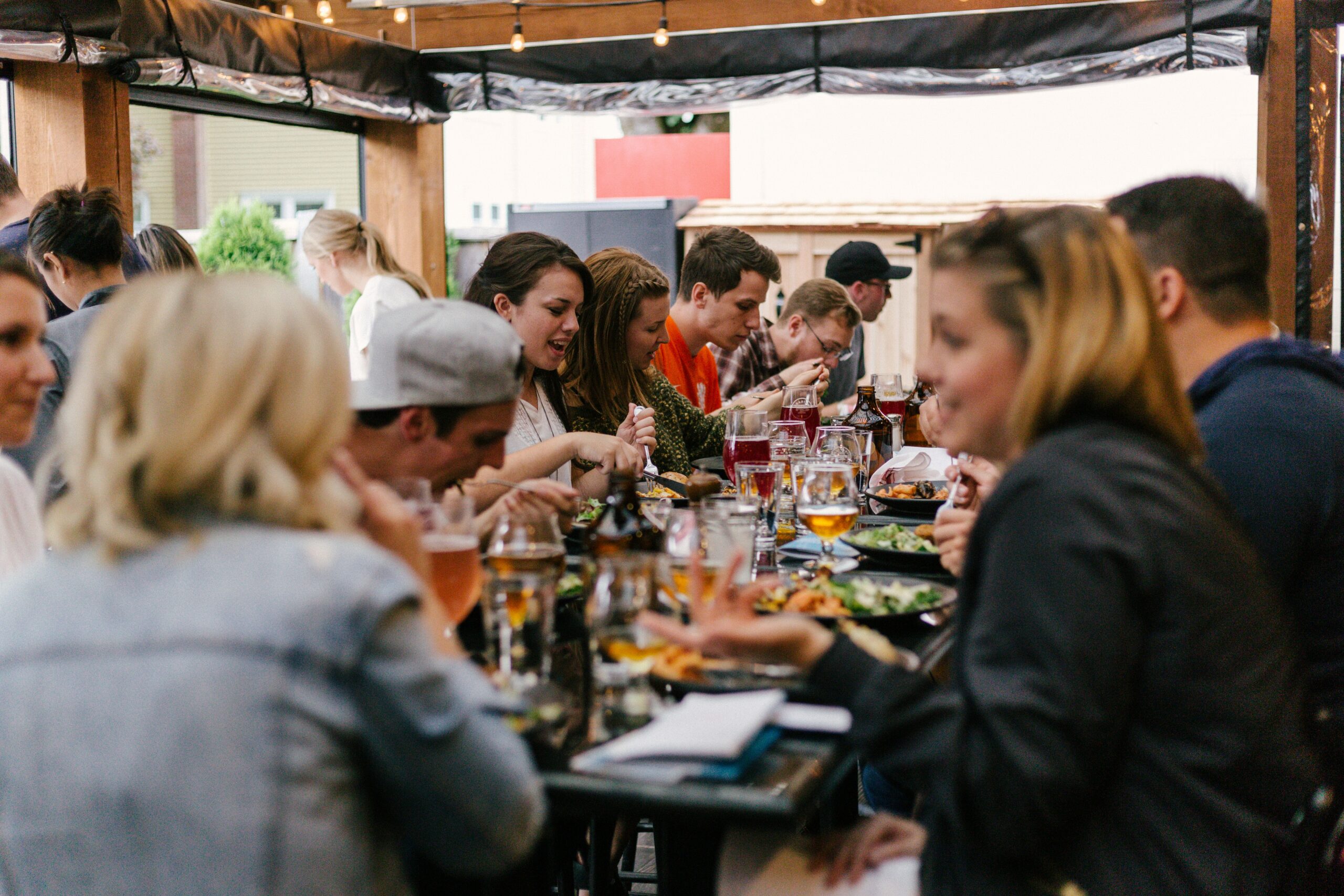 Group of diverse people enjoying a meal at a busy outdoor restaurant with glasses of beer and plates of food on a wooden table.