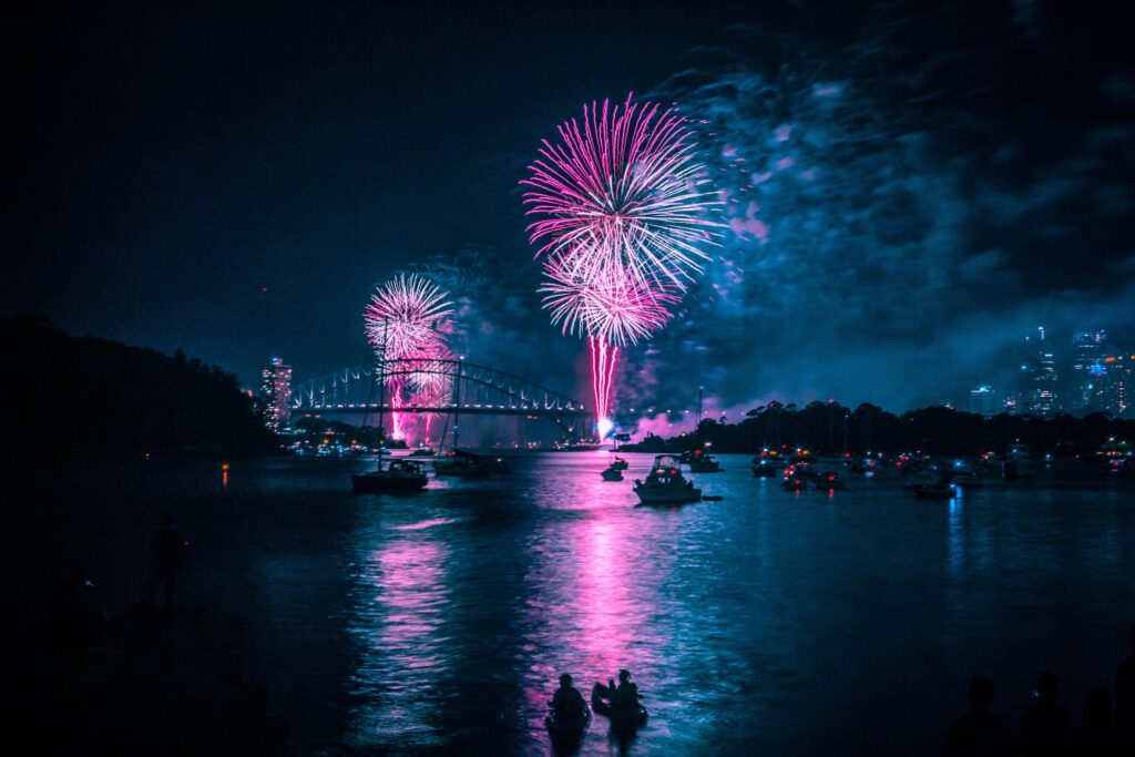 A vibrant fireworks display illuminating the night sky over Sydney Harbour Bridge, with silhouettes of spectators and boats in the foreground.