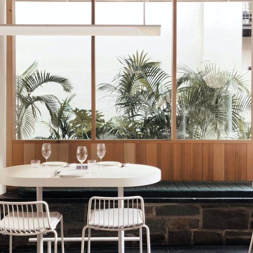 Bright and modern dining area with a white table and chairs, green leather bench, and large windows showcasing tropical plants outside in an Adelaide café.